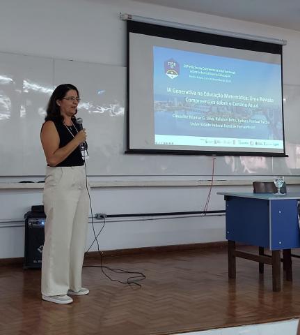 Professora taciana pontual está em pé falando sobre seu trabalho, em frente a uma tela projetada